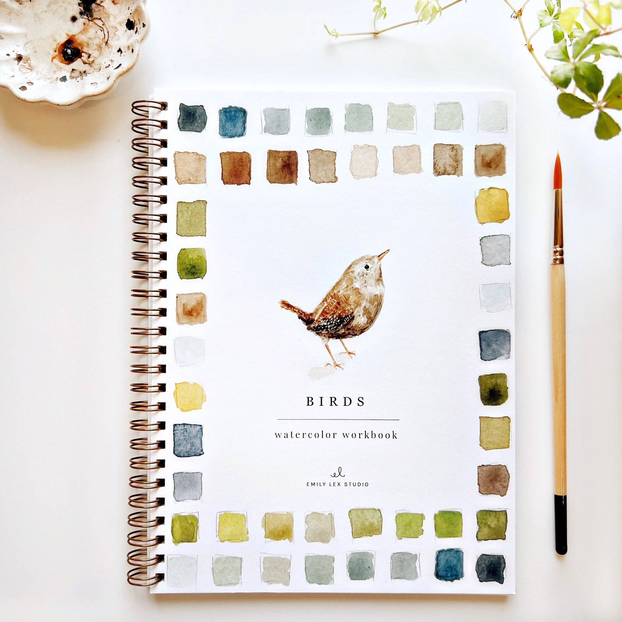 The Faire Watercolor Workbook for beginners, titled "Birds," is spiral-bound and features a painted bird cover with colorful paint swatches. It’s shown on a white surface beside a paintbrush and leafy plant.