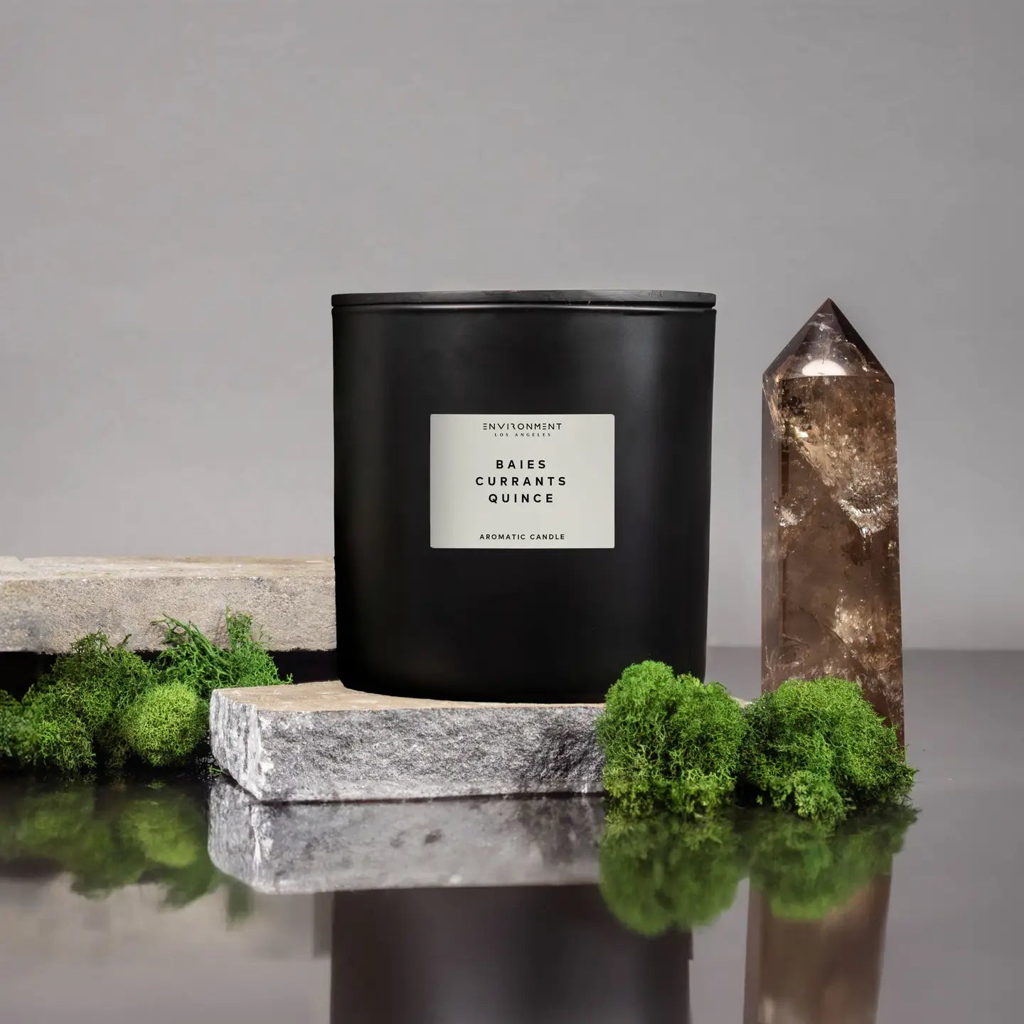 The 55 OZ Diptyque Candle by Faire, crafted from coconut wax, stands on a stone slab. Its black pepper scent blends with earthy green moss beside a brown crystal tower, all set against a gray background.