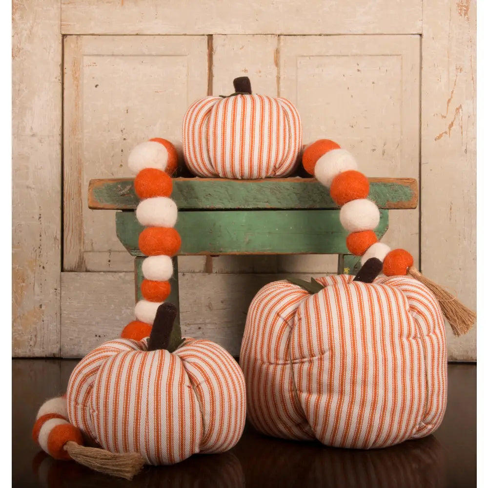 Three Faire Pumpkin decor pieces with orange and white stripes are paired with a felt ball garland on a small green bench, creating charming farmhouse style against a rustic wood background.