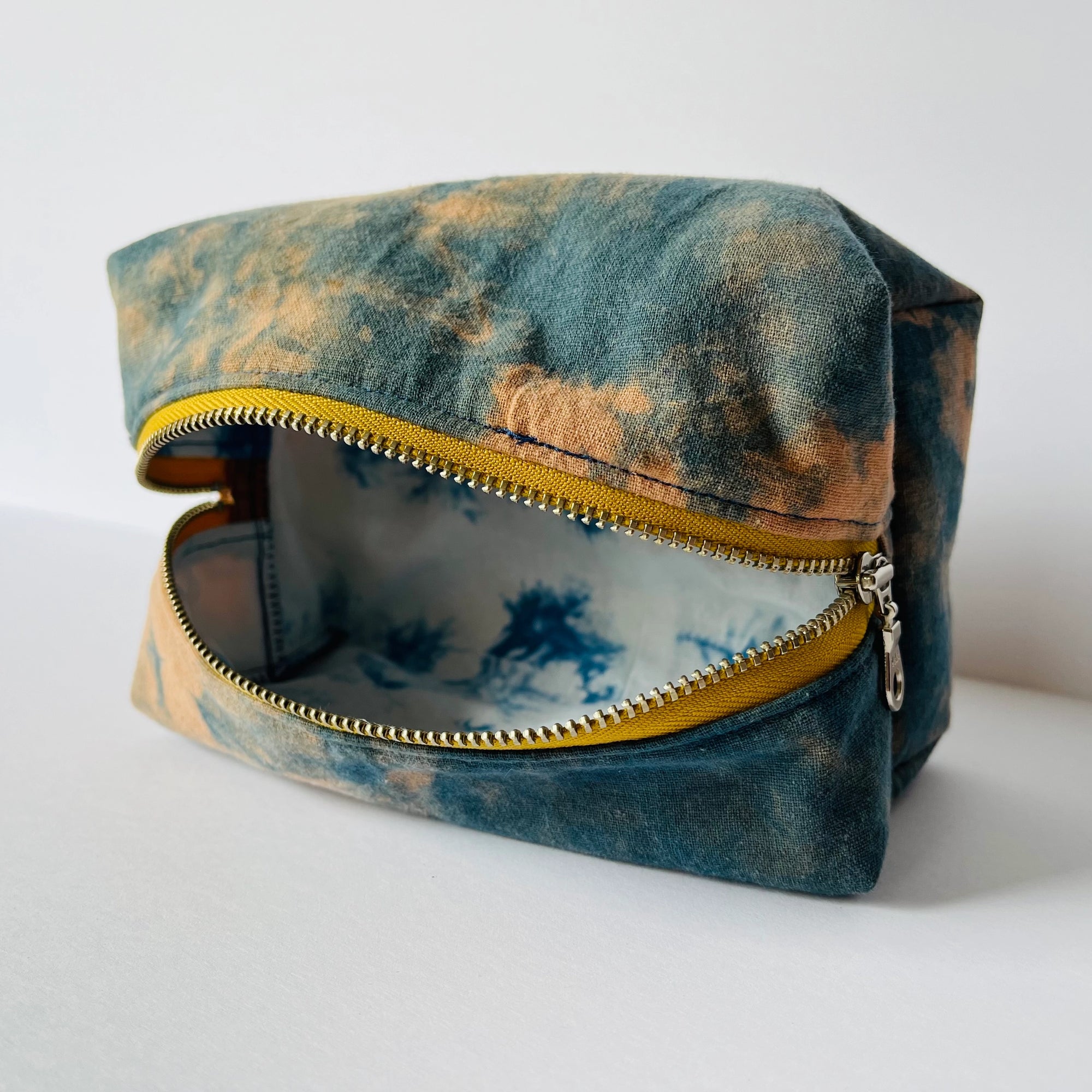 The Faire Dyed Toiletries Bag is a rectangular cotton pouch with a yellow zipper, featuring a blue and tan natural tie-dye exterior and a blue and white tie-dye lining, shown open on a white surface.