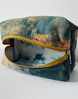 The Faire Dyed Toiletries Bag is a rectangular cotton pouch with a yellow zipper, featuring a blue and tan natural tie-dye exterior and a blue and white tie-dye lining, shown open on a white surface.