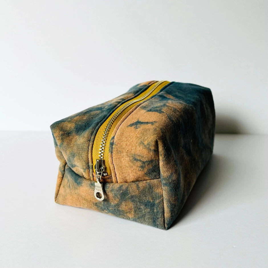The Dyed Toiletries Bag by Faire is a rectangular, hand-dyed cotton pouch with a yellow zipper and blue-brown tie-dye pattern—ideal for organizing your desk.