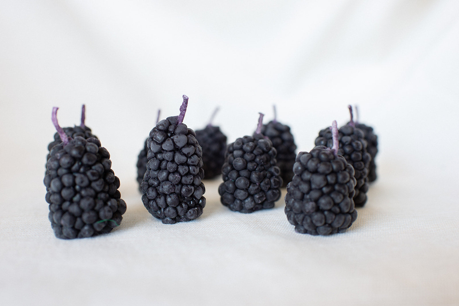 Eight Faire Blackberry Beeswax Candles stand upright on a soft, light surface. Each candle features a purple stem and is spaced unevenly in two loose rows with a white background.