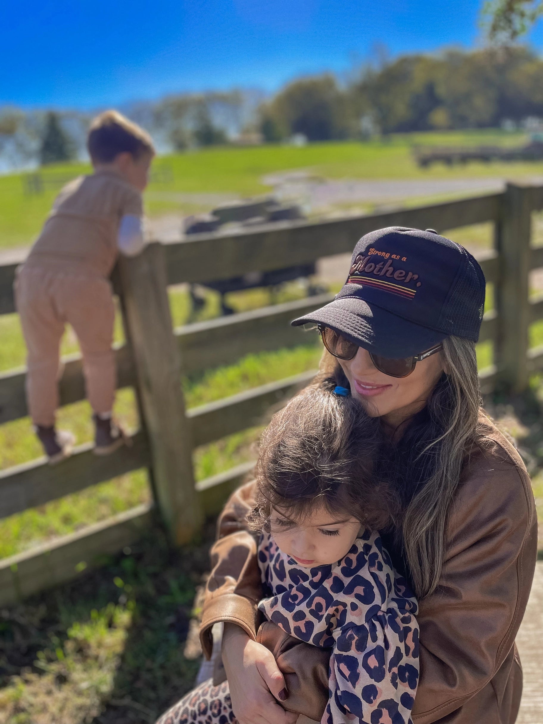 A woman in sunglasses and a Retro Mother Hat by Faire holds a child on her lap, both dressed warmly outdoors. Another child climbs a wooden fence nearby on a sunny day with green grass and trees in the background.