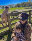 A woman in sunglasses and a Retro Mother Hat by Faire holds a child on her lap, both dressed warmly outdoors. Another child climbs a wooden fence nearby on a sunny day with green grass and trees in the background.