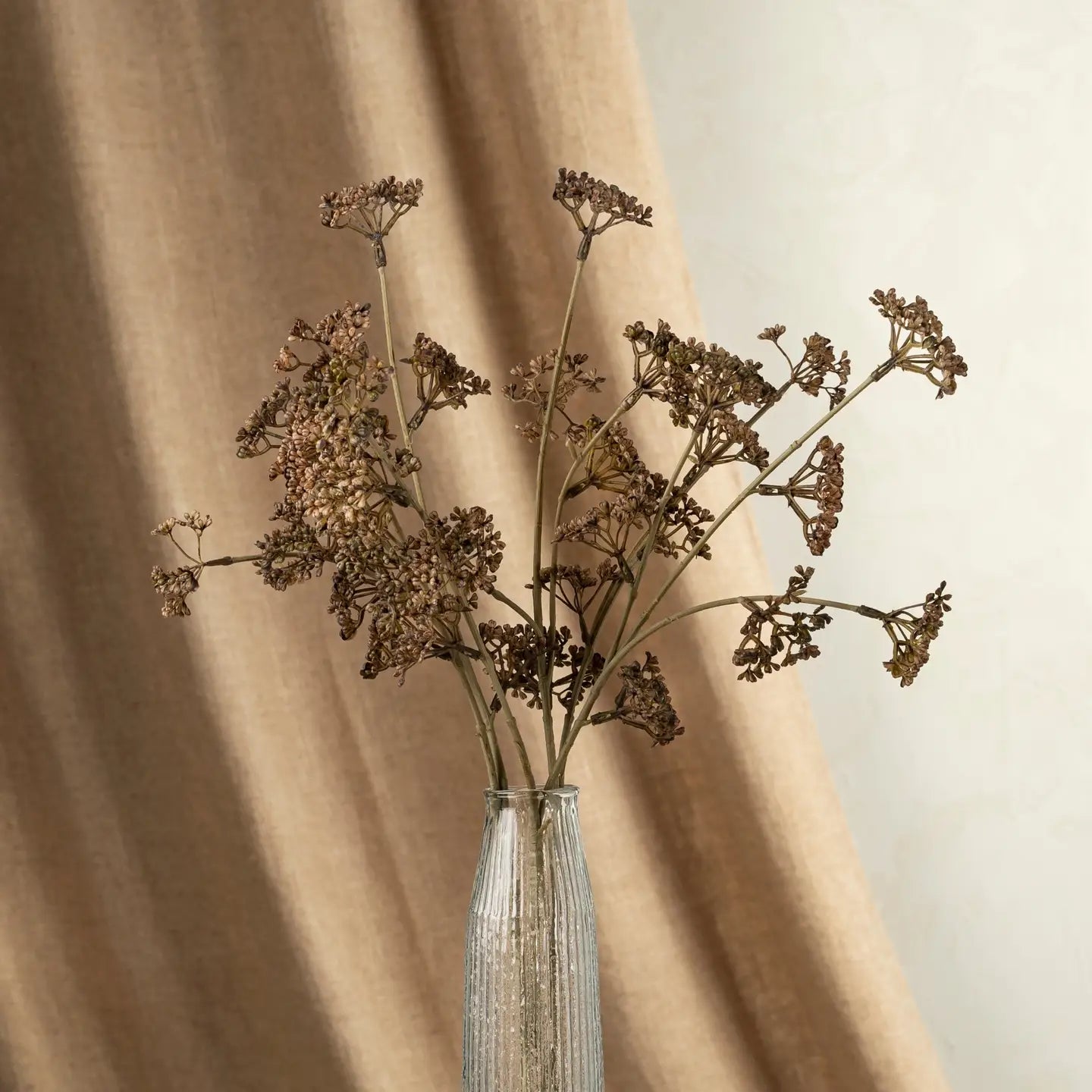 The Indaba Meadow Grass Bouquet, with dried brown stems and clustered flowers, sits in a clear glass vase against a beige fabric and off-white wall—offering natural faux botanical decor.