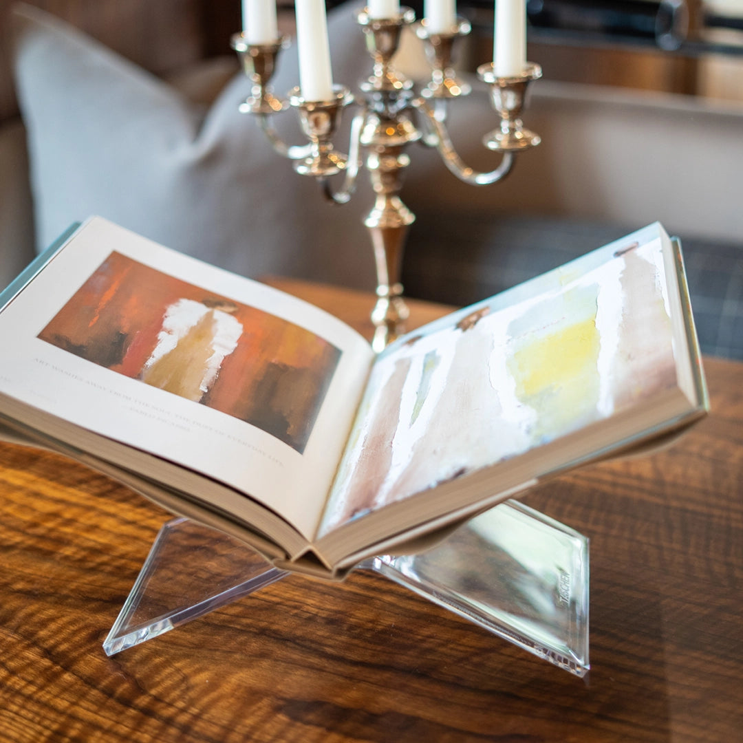 An open coffee table book is showcased on the Faire Acrylic Book Stand Display atop a wooden table next to a silver candelabra, with a blurred pillow and plaid furniture in the background.