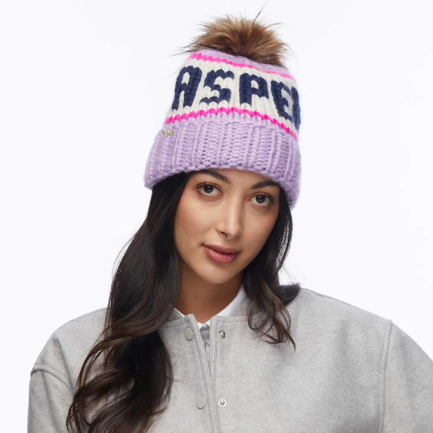 A woman with long dark hair wears the Faire Knit Ski Hat, a light purple hand-knit beanie with a brown faux fur pom pom and "ASPEN" lettering. She pairs it with a light gray jacket and looks at the camera against a plain white background.