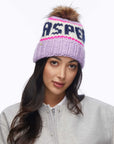 A woman with long dark hair wears the Faire Knit Ski Hat, a light purple hand-knit beanie with a brown faux fur pom pom and "ASPEN" lettering. She pairs it with a light gray jacket and looks at the camera against a plain white background.