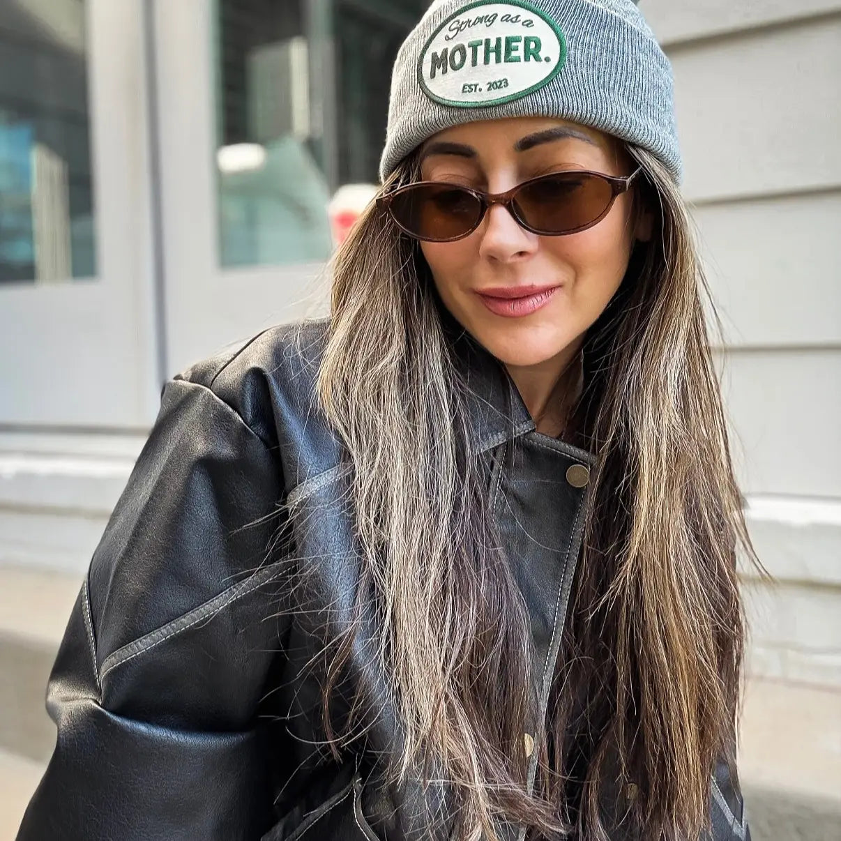 A woman with long, light brown hair smiles softly while wearing the Faire Patch Beanie Strong as a Mother in gray, paired with brown sunglasses and a black leather jacket outdoors in front of a building.