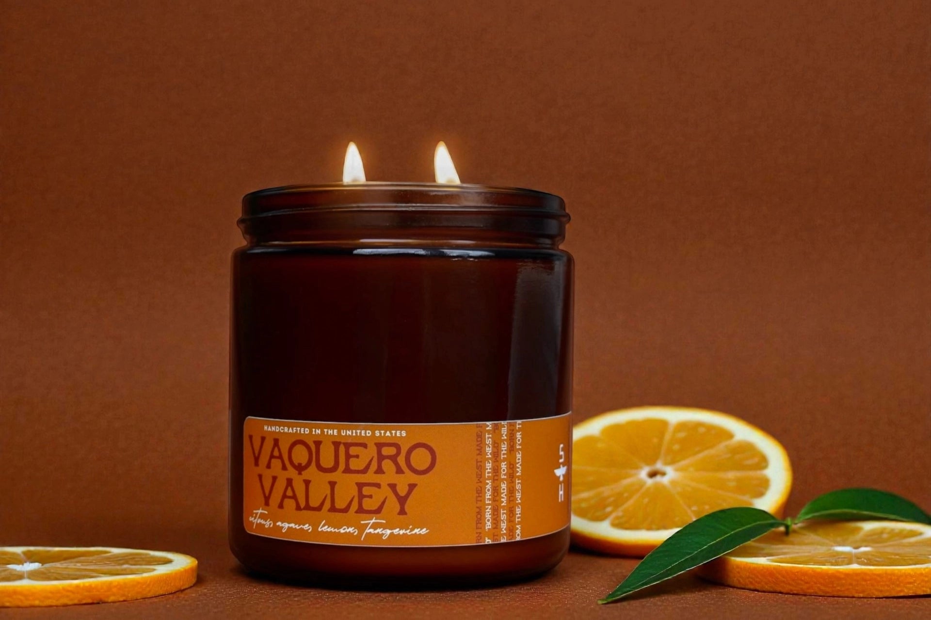 A brown glass Faire Citrus and Agave Candle with two wicks sits on a brown surface, surrounded by orange slices and green leaves. Its label notes a sweet citrus blend of ripe orange, lemon, and tangerine—a Volcano-inspired scent.
