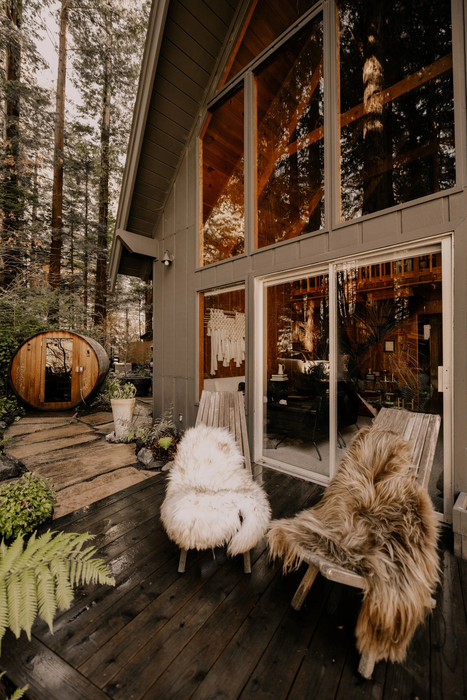 A cozy cabin with large glass windows sits in a forest. On the deck, two wooden chairs feature Faire Icelandic Sheepskin throws, and a wooden barrel sauna stands among tall trees in the background.