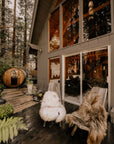 A cozy cabin with large glass windows sits in a forest. On the deck, two wooden chairs feature Faire Icelandic Sheepskin throws, and a wooden barrel sauna stands among tall trees in the background.