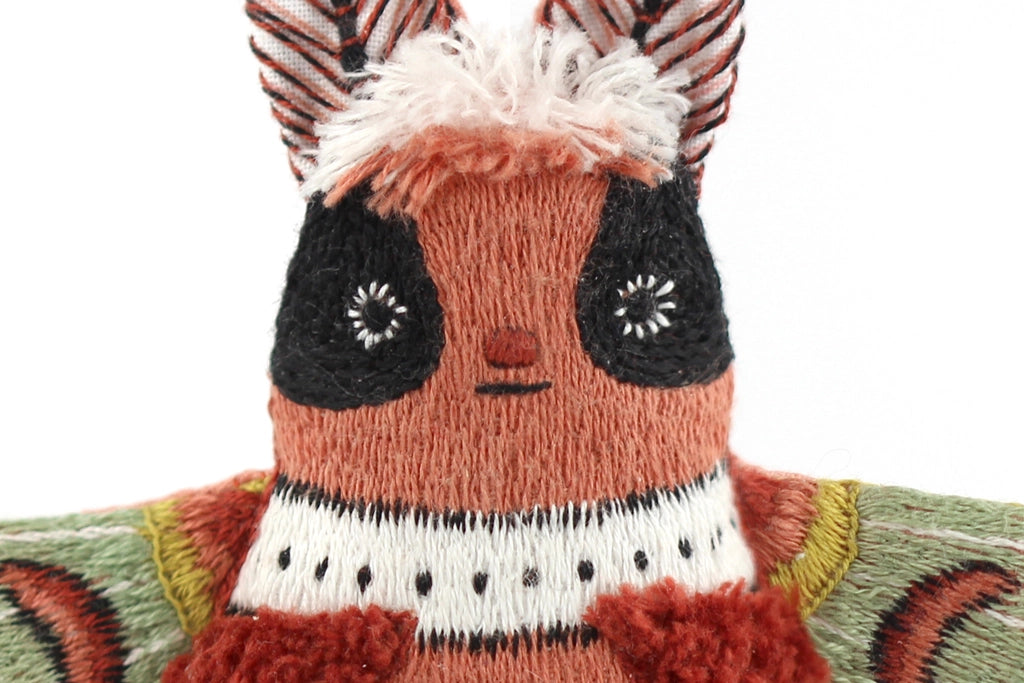 Close-up of a handmade plush moth crafted with the Faire Moth Embroidery Kit, showcasing embroidered facial features, large black eyes, striped antennae, and green wings adorned with red and black accents.