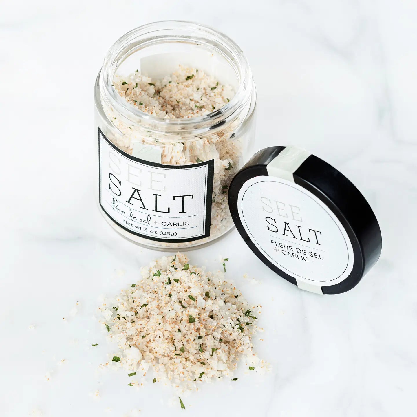 An open jar of Faire's Fleur de Sel Sea Salt is shown with coarse salt and herbs spilled on a white surface. The black lid with the product label lies next to it.