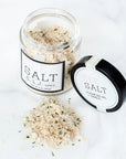 An open jar of Faire's Fleur de Sel Sea Salt is shown with coarse salt and herbs spilled on a white surface. The black lid with the product label lies next to it.