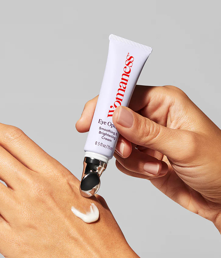 A hand dispenses Faire Eye Opener - Smoothing & Brightening Cream, a white formula with a metal applicator tip, onto the back of another hand—ideal for puffiness and dark circles—all set against a neutral background.