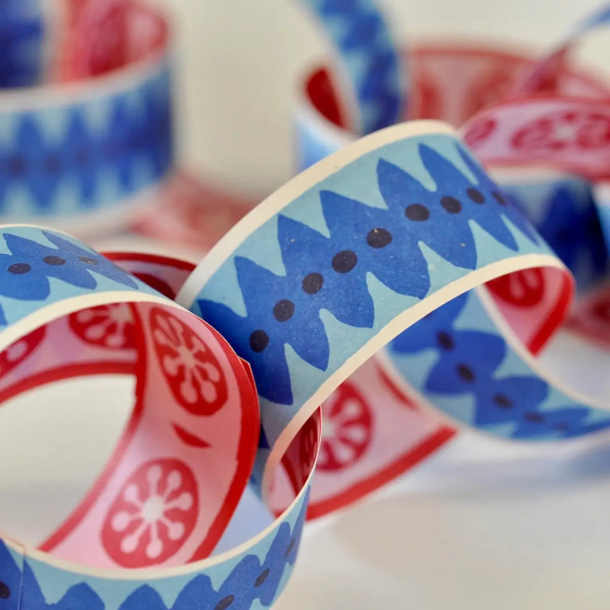 Faire's Paperchain features colorful, biodegradable decorative links with blue, red, and pink geometric and floral patterns, arranged loosely overlapping on a light surface.