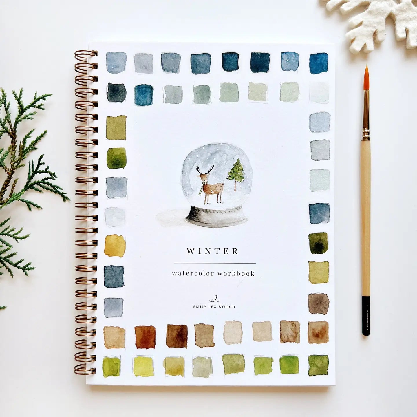 The spiral-bound Faire Watercolor Workbook, titled &quot;Winter,&quot; showcases a snow globe with a deer, trees, and color swatches on the cover. Beside it are a pine sprig, paintbrush, and snowflake ornament set against a white background.