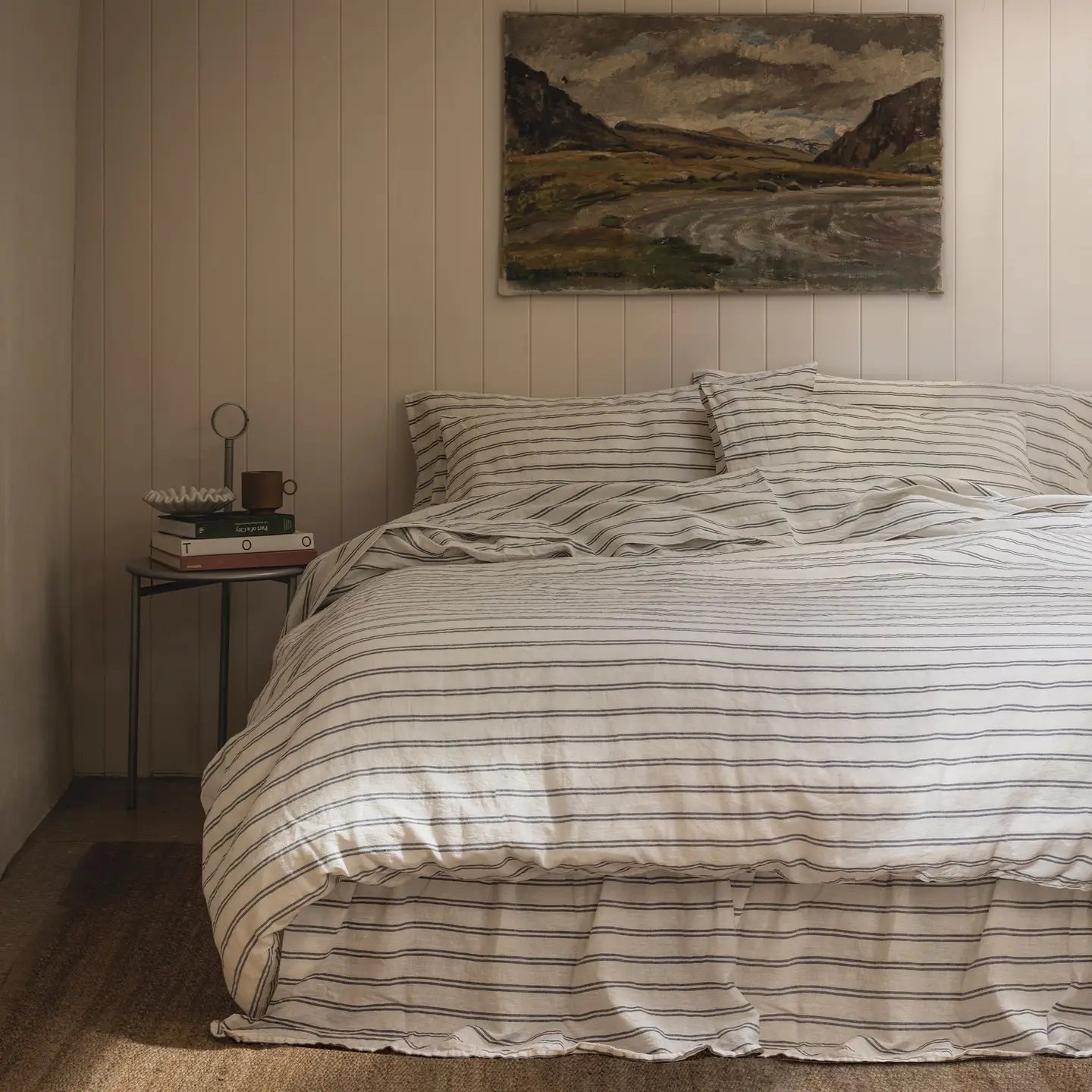 A neatly made bed features the Faire Kemptown Stripe Linen Duvet against a beige paneled wall. A landscape painting hangs above, while a bedside table holds books, a lamp, and decorative decor.