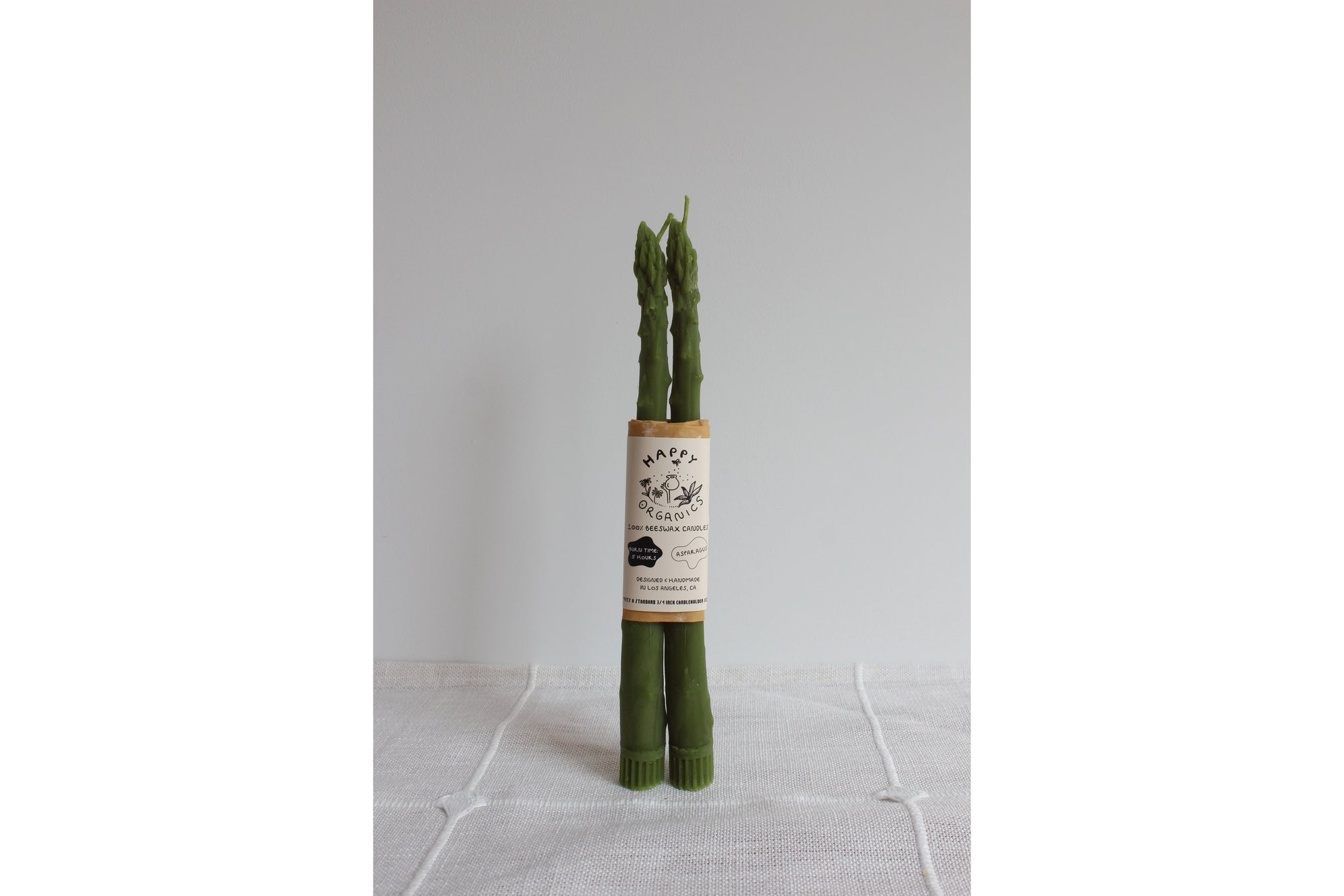 Two upright asparagus stalks, appearing lifelike, are actually Faire's Asparagus Beeswax Candles. Handmade with beeswax, they stand on a plain white cloth and are bound together by a label against a simple light background.