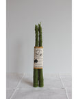Two upright asparagus stalks, appearing lifelike, are actually Faire's Asparagus Beeswax Candles. Handmade with beeswax, they stand on a plain white cloth and are bound together by a label against a simple light background.