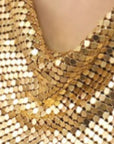 A close-up of shimmering gold sequin fabric in folds, showcasing Faire's Chain Mail Bandana Scarf Necklace at the neckline.