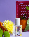 Maison Matine Perfume by Faire—a clear bottle with a white cap and colorful cartoon designs—rests on a purple surface with orange and yellow flowers, green chrysanthemum buds, tangerine, and hints of vanilla against a purple background.