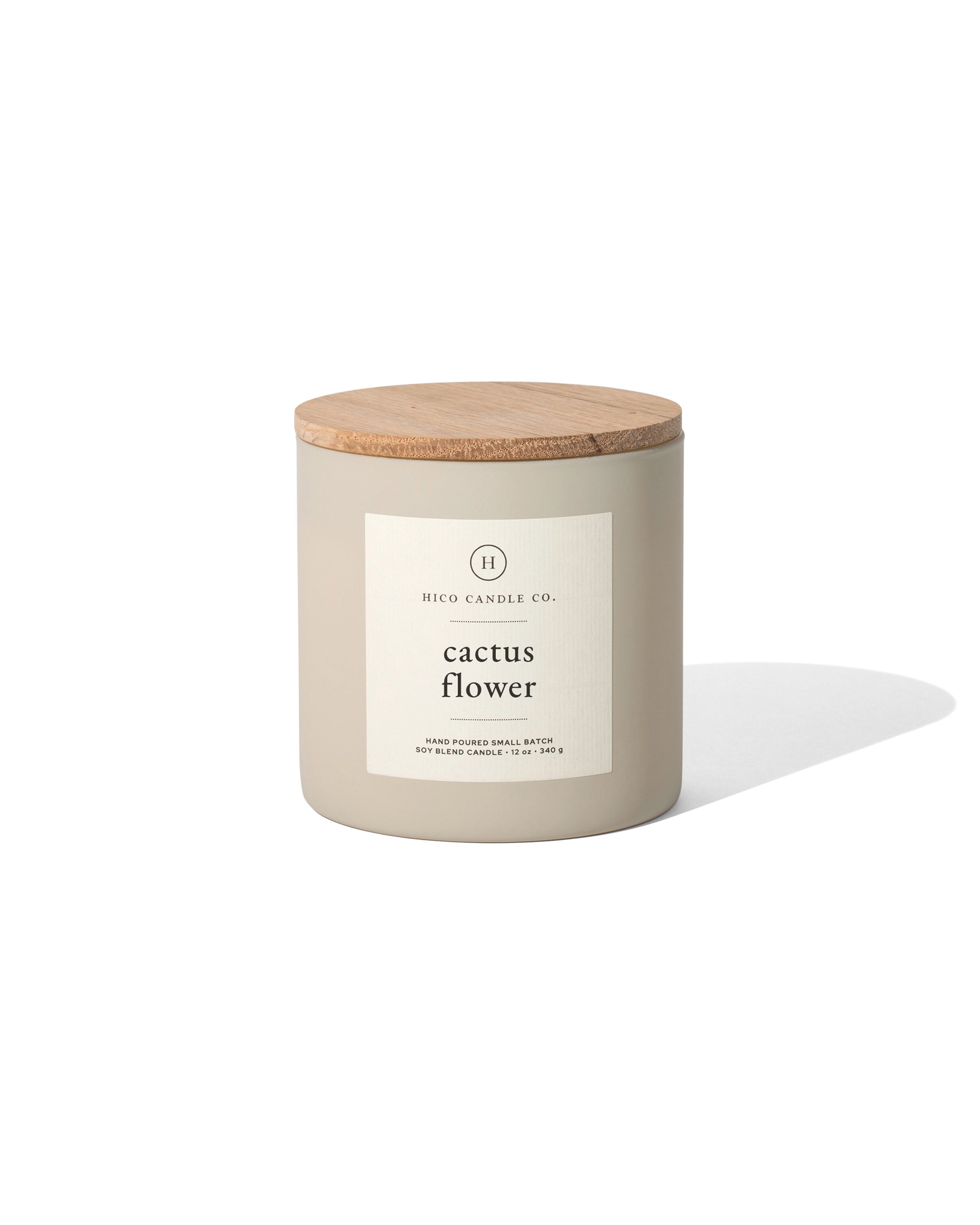 A beige 14oz. soy wax candle from Faire with a wooden lid and white label reading "Cactus Flower" sits on a white background, casting a soft shadow to the right. It offers a delightful floral fragrance.