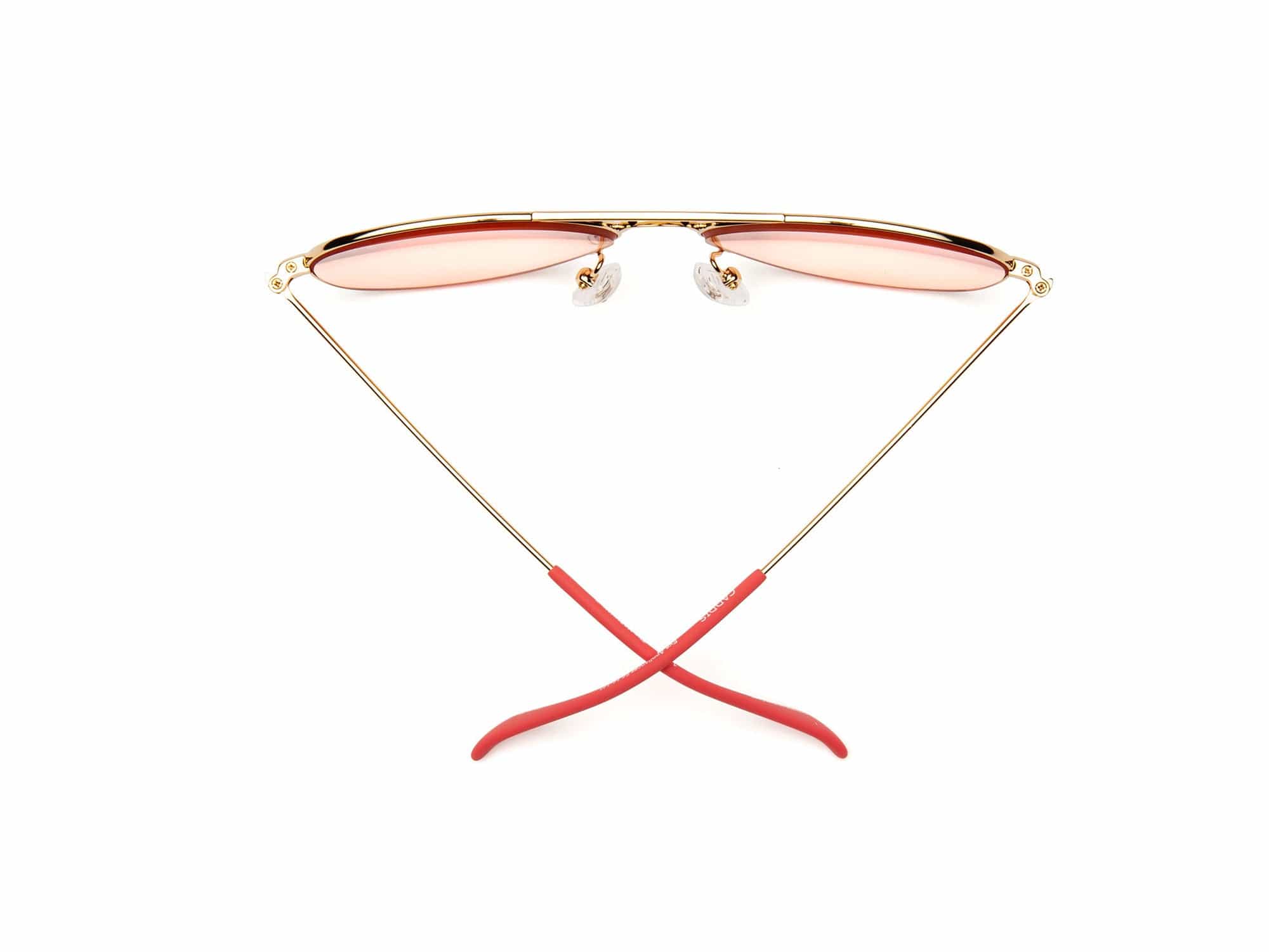 Caddis Mabuhay Polished Gold aviator sunglasses with gold wire frames, pink-tinted lenses, and red rubber temple tips are displayed upright with crossed arms on a white background.