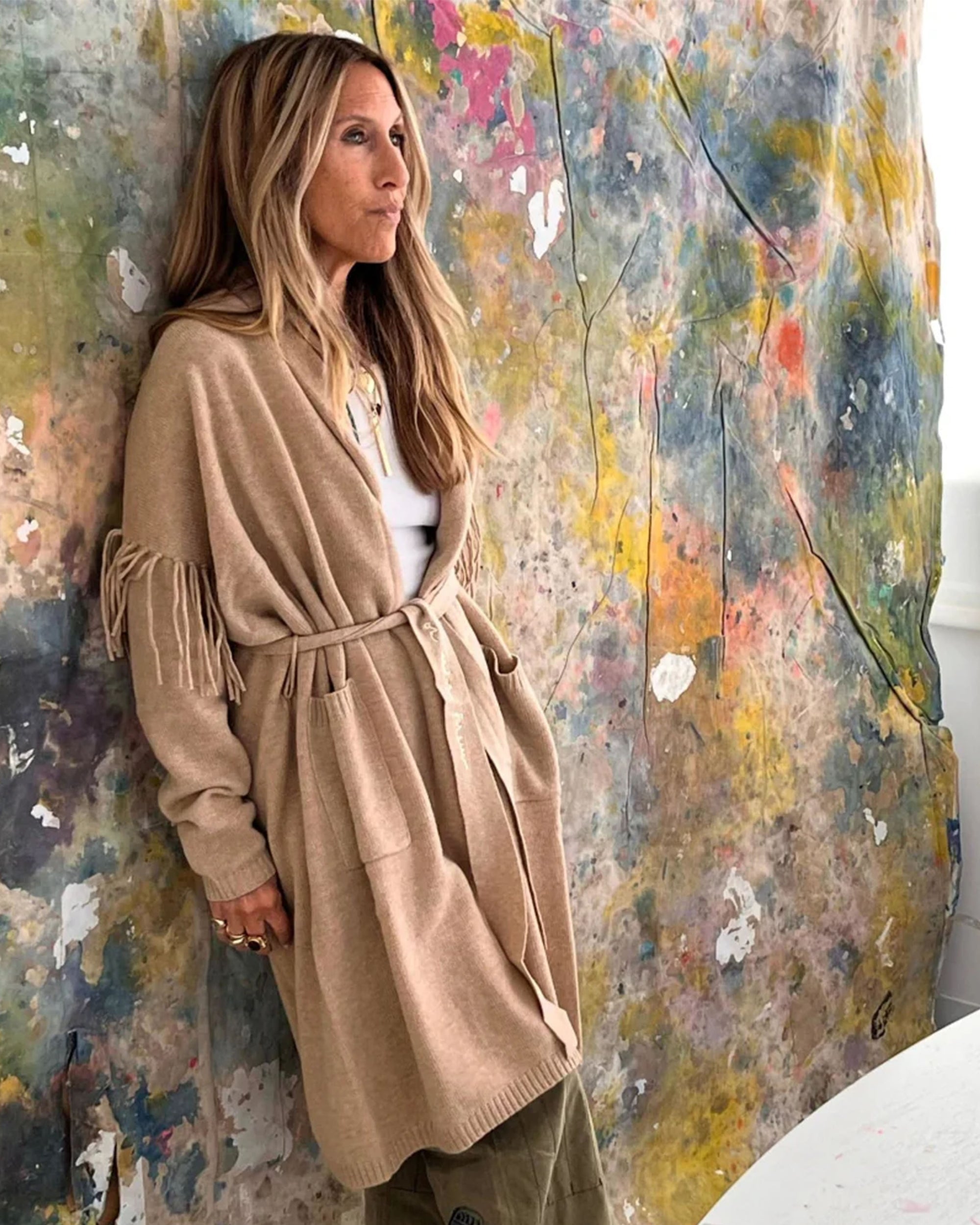 A woman with long hair wears the Kerri Rosenthal Cali Fringe Cardigan Coat, paired with green pants, standing against a colorful, abstract painted backdrop and gazing thoughtfully to the side.