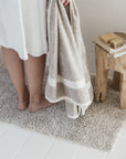 A person in a white towel stands barefoot on a plush, beige Calisto Bath Rug by Libeco Home. They hold a large, beige towel with white stripes, made from a premium linen-cotton blend. Next to them is a small wooden stool with a folded towel and bar of soap on top, against a light-colored wall and white floor.