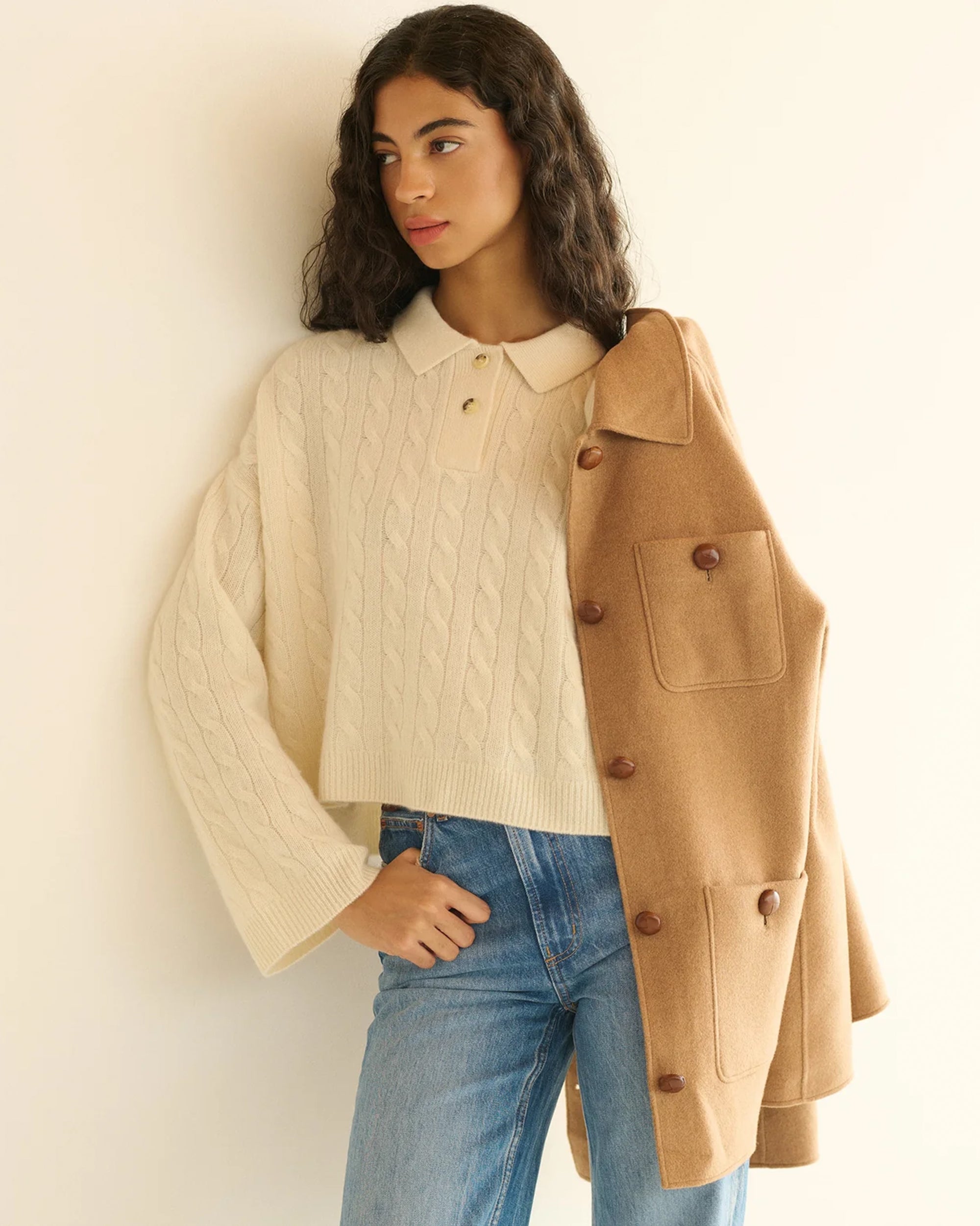 A woman with long curly hair wears the DOEN Cambridge Cashmere Sweater in cream, light blue jeans, and drapes a camel-colored coat over her shoulder, standing against a neutral background.