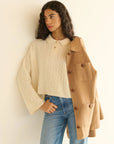 A woman with long curly hair wears the DOEN Cambridge Cashmere Sweater in cream, light blue jeans, and drapes a camel-colored coat over her shoulder, standing against a neutral background.