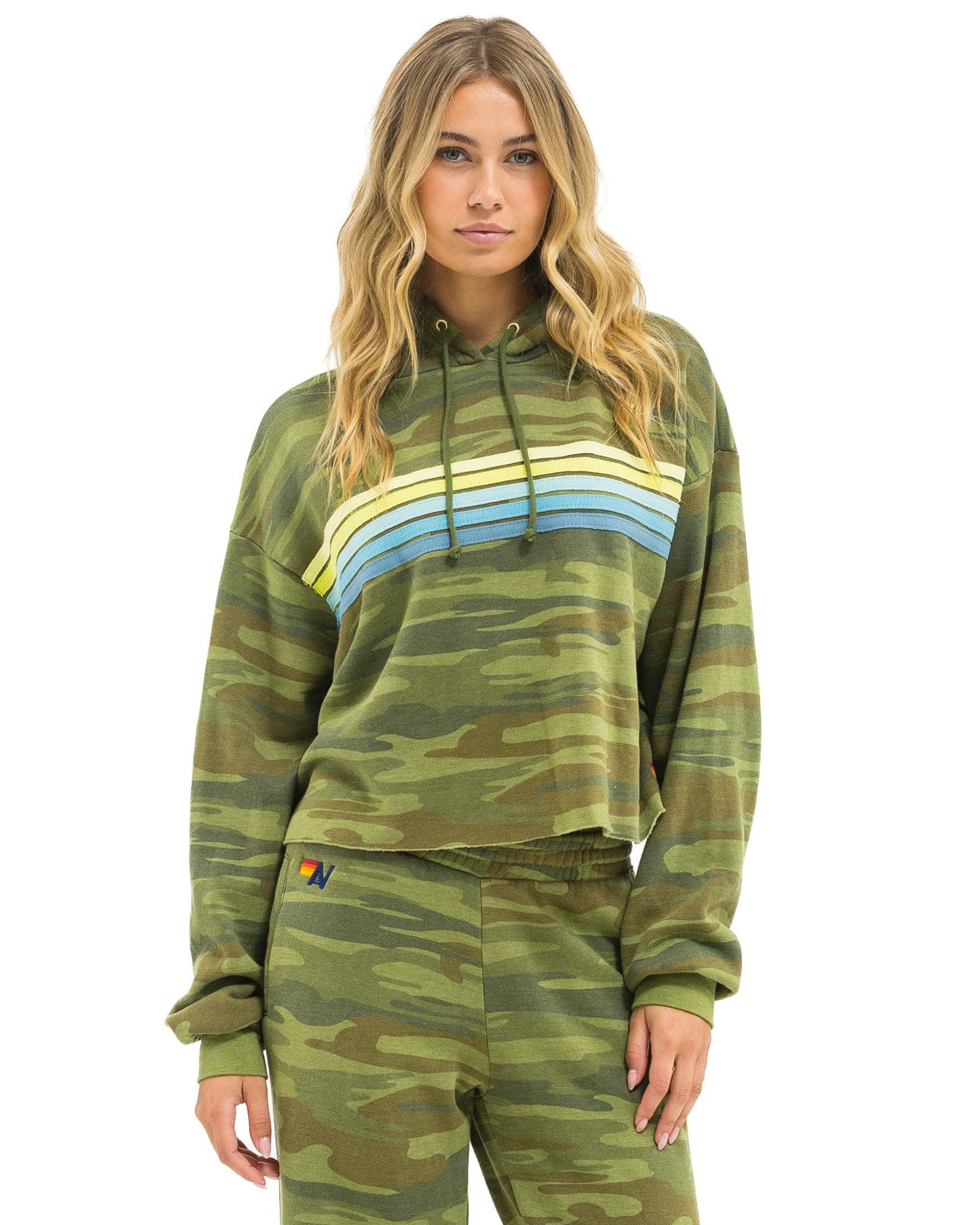 A woman with long, wavy blonde hair wears the Aviator Nation 5 Stripe cropped pullover hoodie relaxed in green camo with rainbow stripes and matching pants, posing against a white background for a cool vintage vibe.