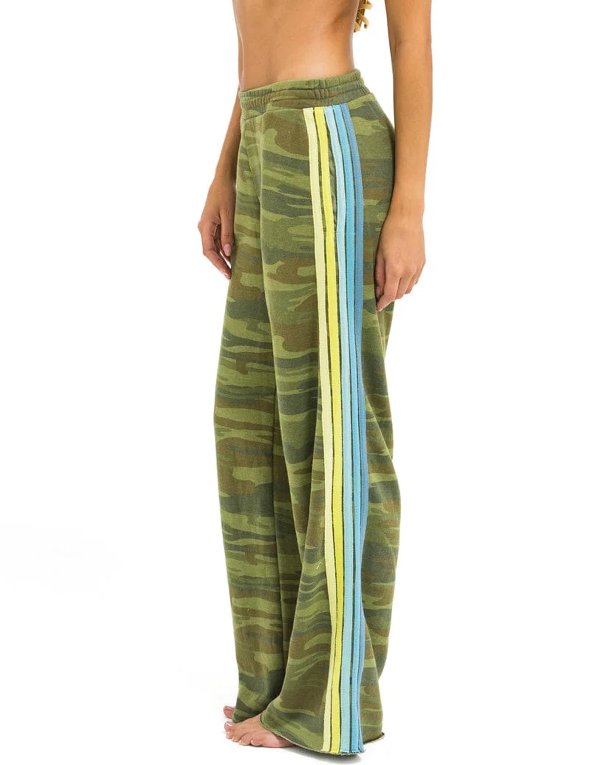 A person wears Aviator Nation's 5 stripe women's wide leg pocket sweatpants—vintage, loose-fitting green camo with blue and yellow vertical side stripes—standing barefoot against a plain white background.