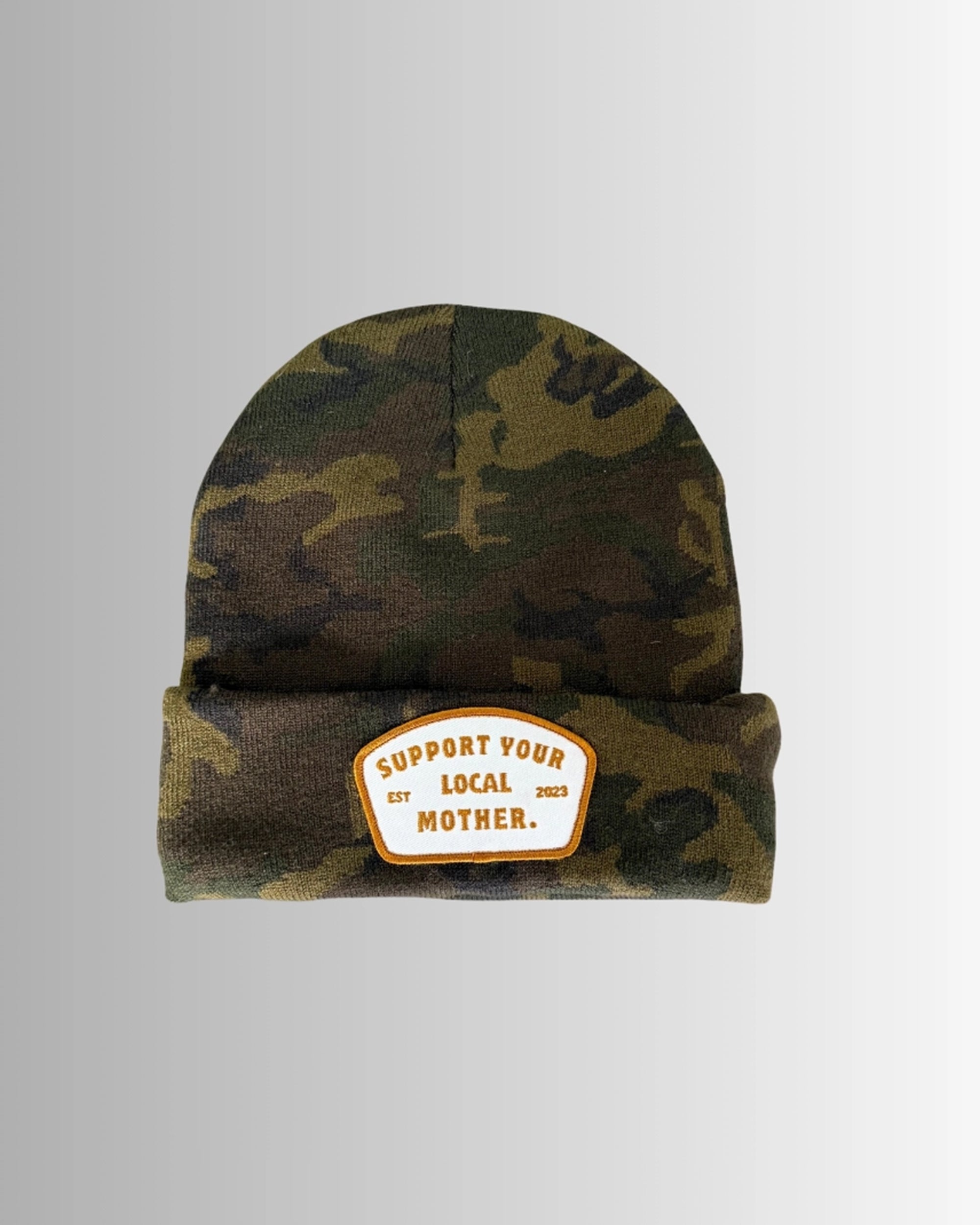 The Faire Patch Beanie Support Your Local Mother features a camo knit, folded brim, and a white-orange patch with "SUPPORT YOUR LOCAL MOTHER. EST 2023." Displayed on a light gradient background.