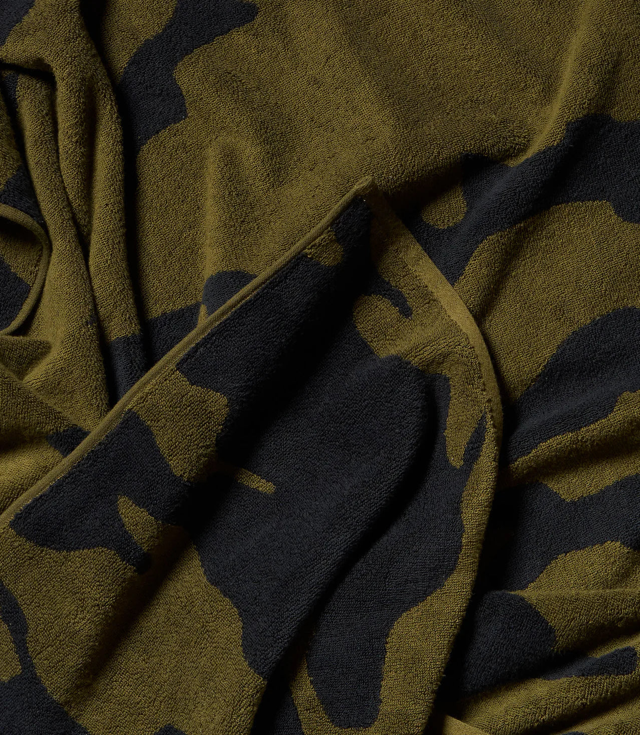 A close-up of the Typical Wild Camouflage Bath Towel highlights its soft, textured green and black design with overlapping folds and subtle shadows.