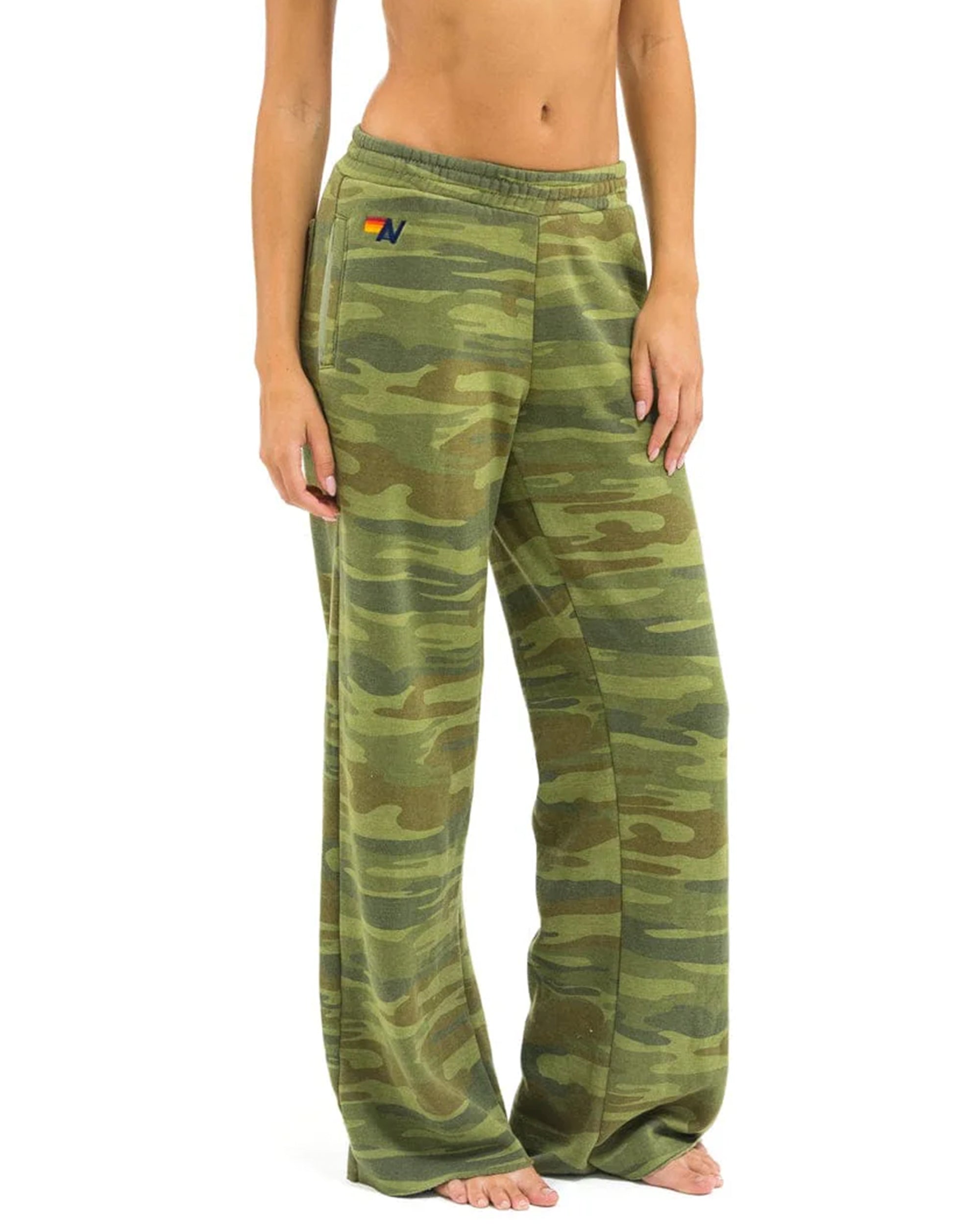 A person stands barefoot in loose, green camouflage Aviator Nation 5 Stripe women’s wide leg pocket sweatpants with an elastic waistband and a small embroidered logo by the left pocket. The image is cropped from shoulders to feet.
