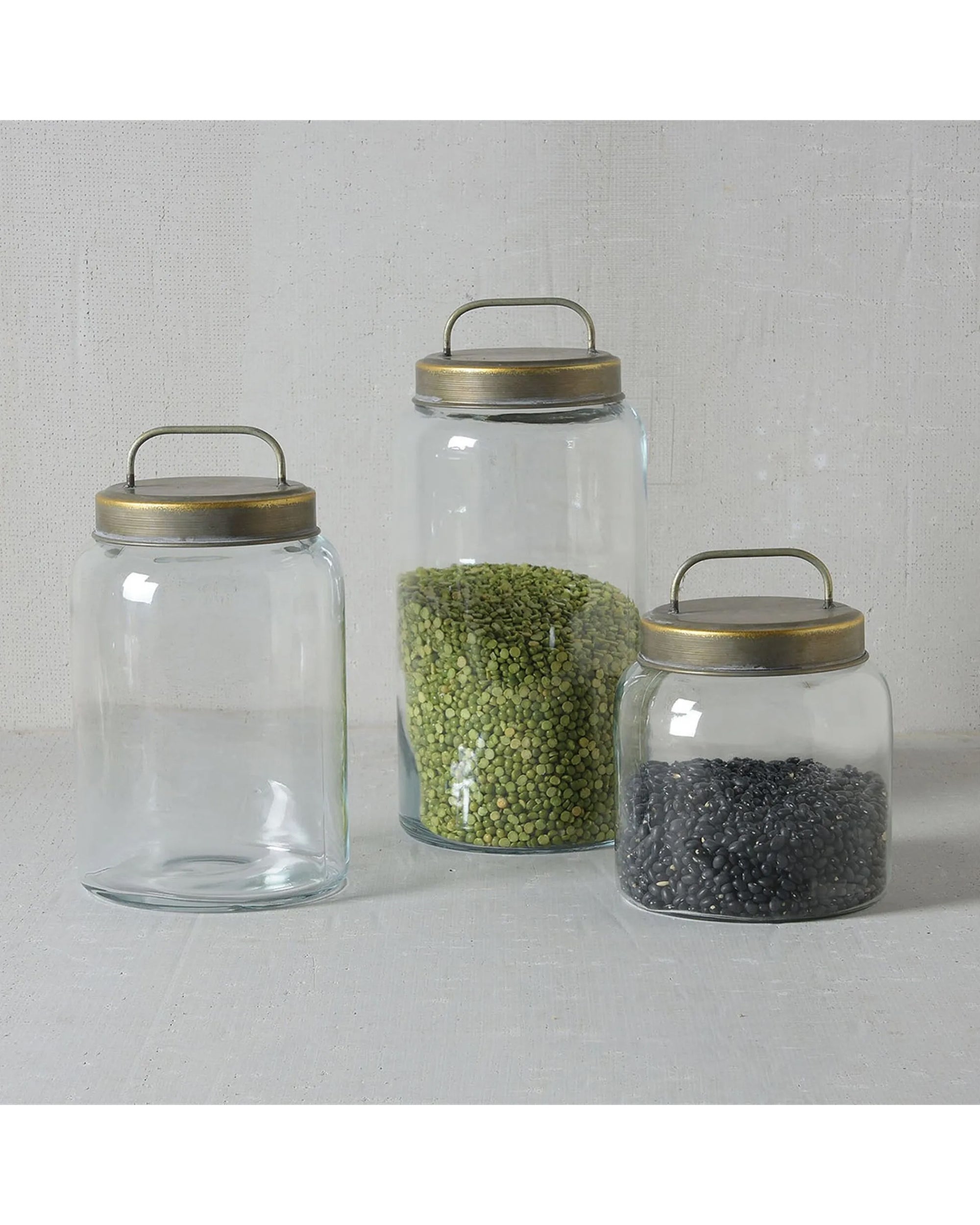 Three HomArt 50011/2/3 Archer Canister Metal Lid jars are shown: the tallest is half-filled with green lentils, the shortest holds black beans, and the medium jar is empty. All are placed on a light-colored surface.
