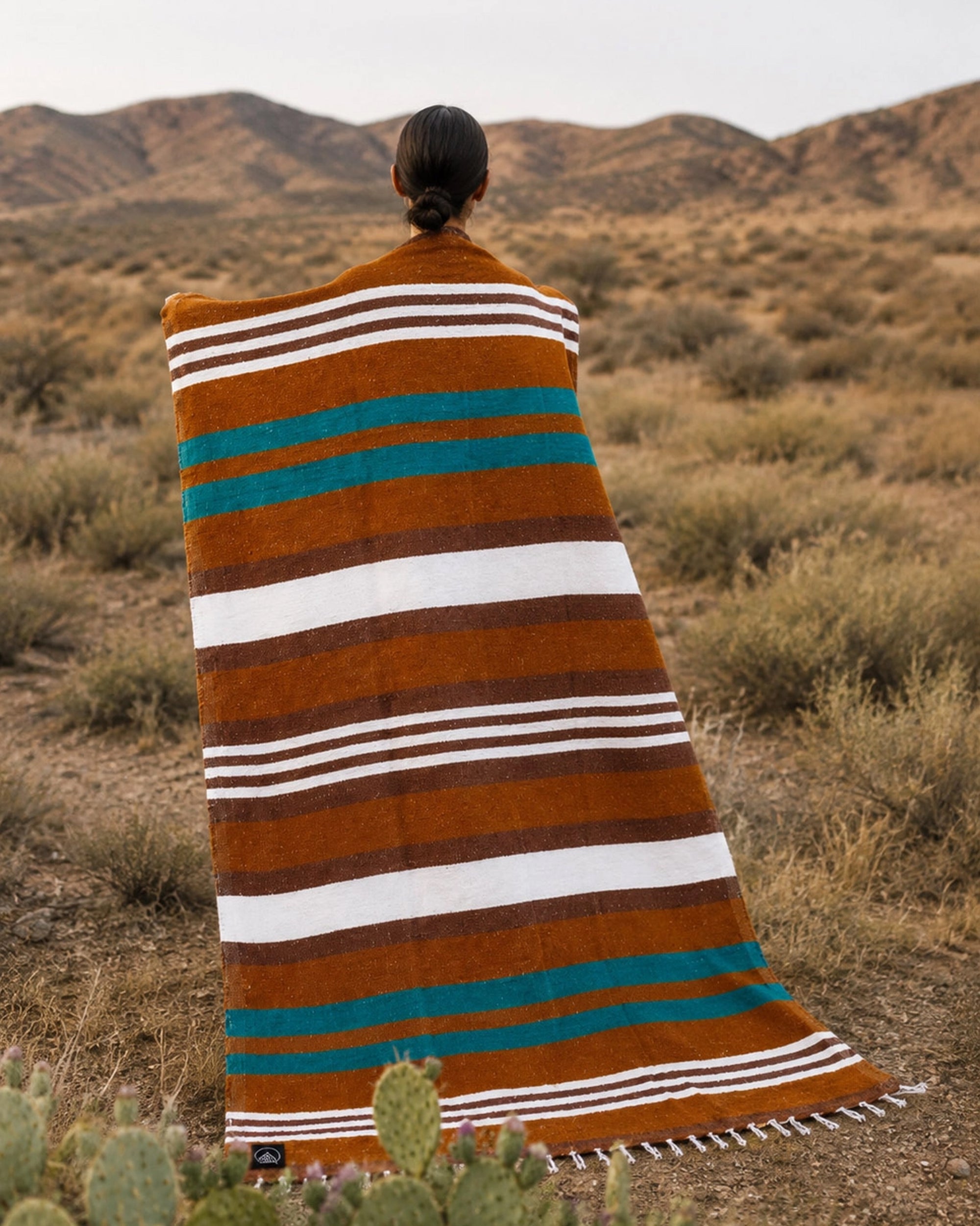 Faire Canyon Cove Handwoven Blanket by Bungalow draped on person in a desert landscape with mountains and shrubs.