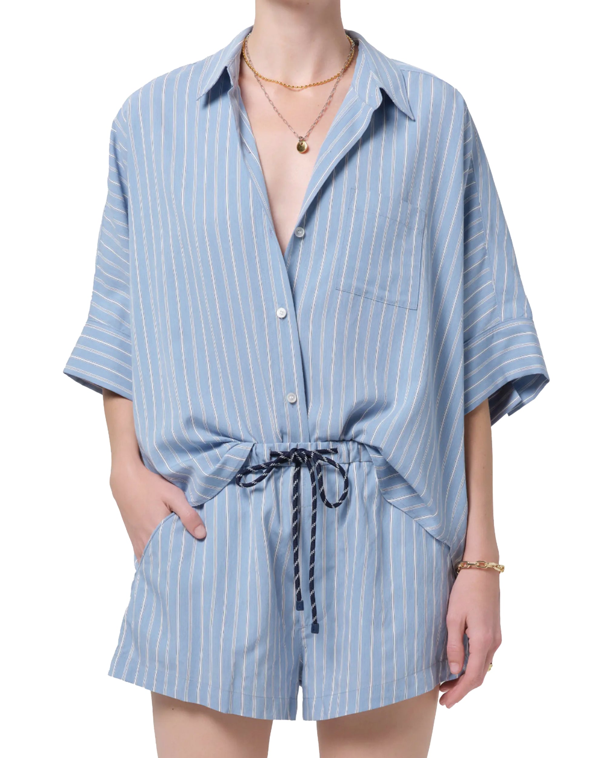 Dressed in the Citizens Of Humanity/AGOLDE Claire Origami Stripe set—a loose, light blue striped button-up shirt and matching drawstring shorts in regenerative cotton—they accessorize with gold jewelry, one hand tucked casually in a pocket.