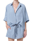 Dressed in the Citizens Of Humanity/AGOLDE Claire Origami Stripe set—a loose, light blue striped button-up shirt and matching drawstring shorts in regenerative cotton—they accessorize with gold jewelry, one hand tucked casually in a pocket.