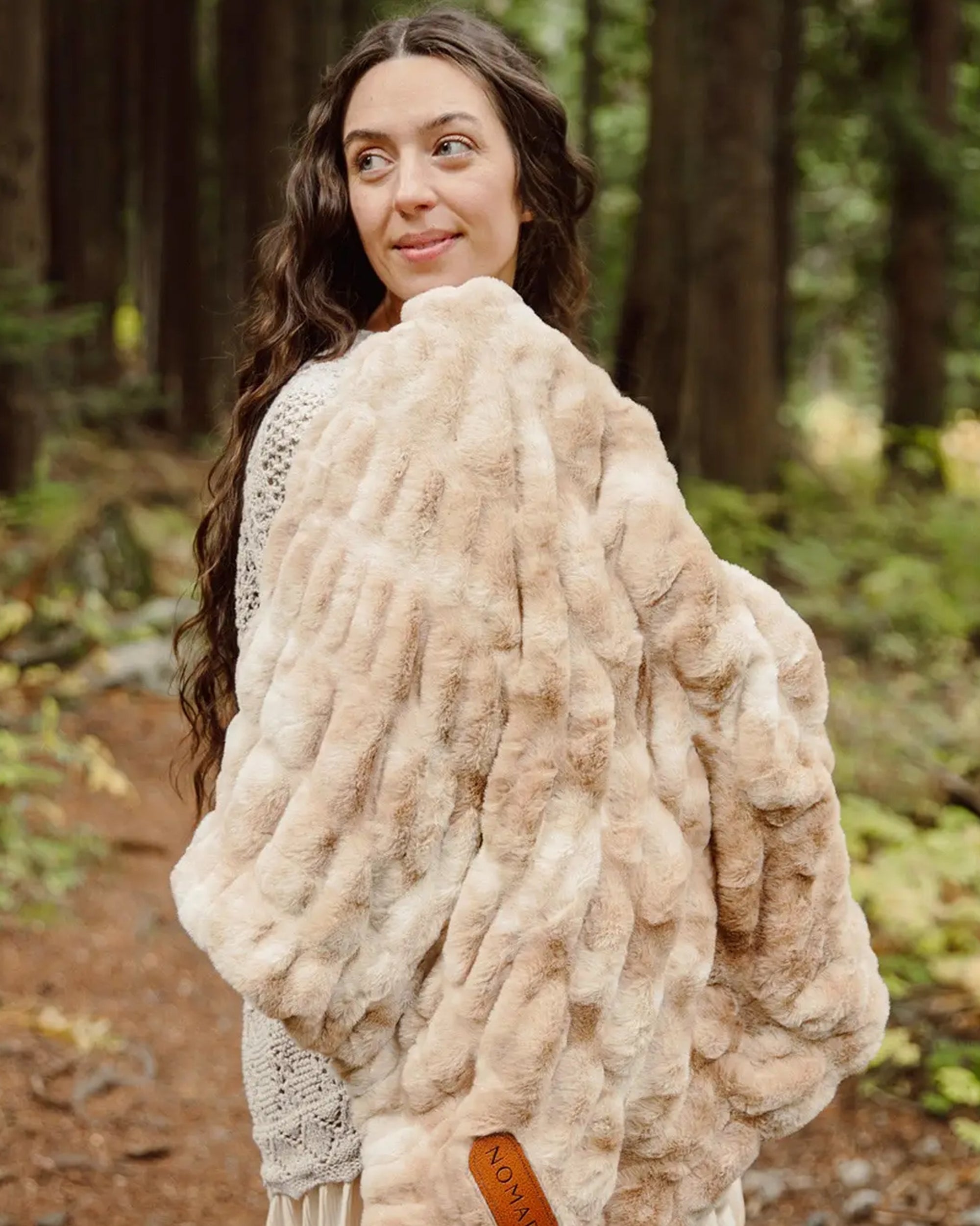 A woman with long wavy brown hair stands in a forest, smiling over her shoulder while wearing the Faire Cascade Throw Blanket and a cream-colored knit sweater.