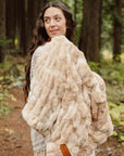 A woman with long wavy brown hair stands in a forest, smiling over her shoulder while wearing the Faire Cascade Throw Blanket and a cream-colored knit sweater.