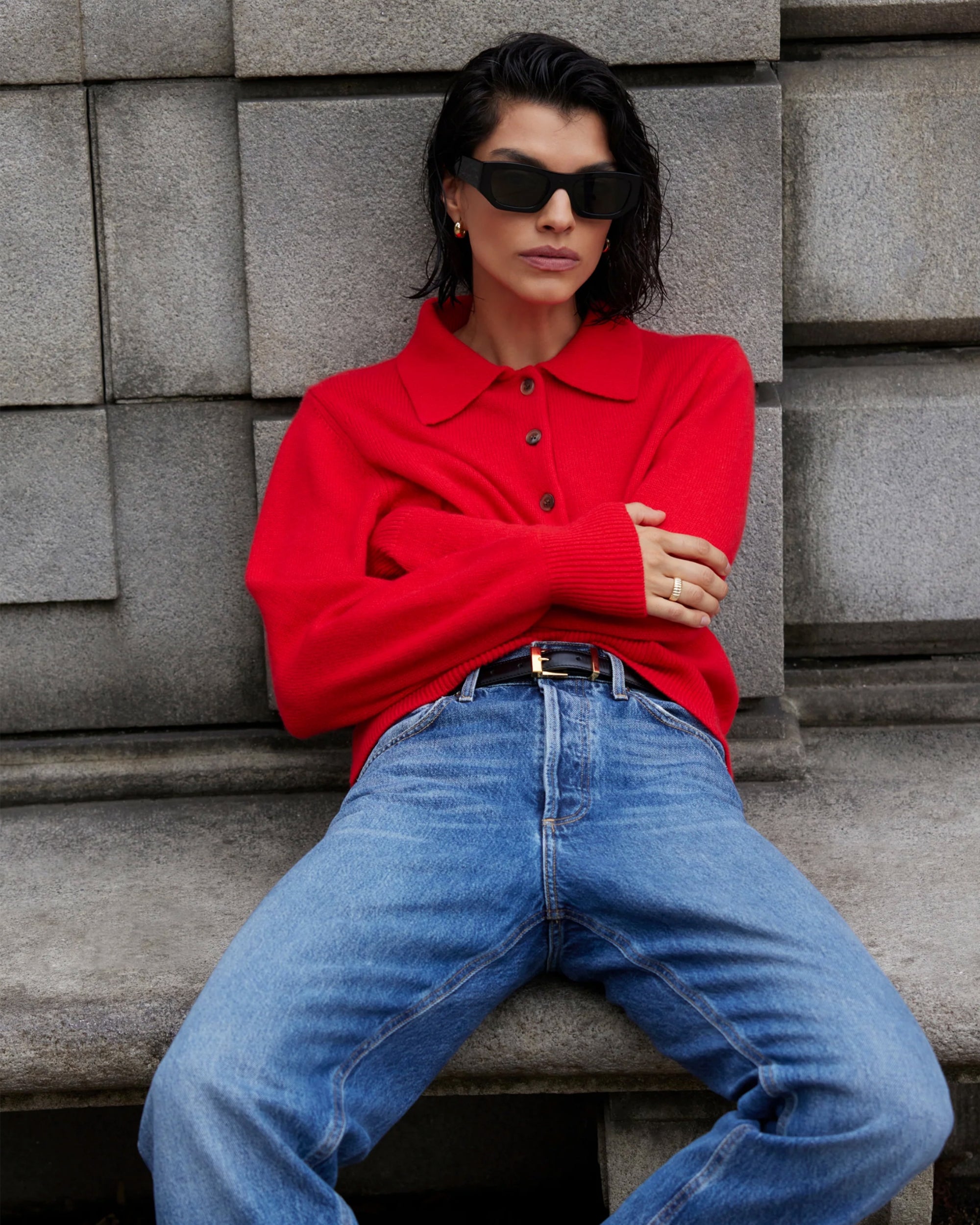 A person with short dark hair, black sunglasses, and blue jeans sits on a stone bench against a gray block wall, arms folded and legs crossed, wearing a bright red Sold Out NYC Cashmere Polo.