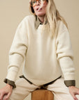 A woman with long blonde hair and sunglasses wears the Sold Out NYC Cashmere Signature Crew in cream over an olive green shirt, seated on a wooden block against a beige background.