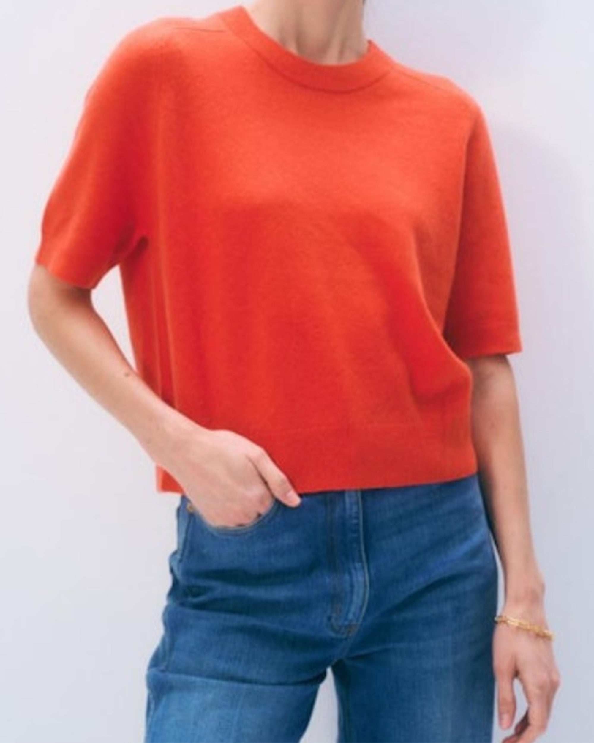 A person wears the White + Warren Cashmere Relaxed T-Shirt in bright orange with blue jeans, standing against a plain light background, one hand in their pocket and the other by their side.