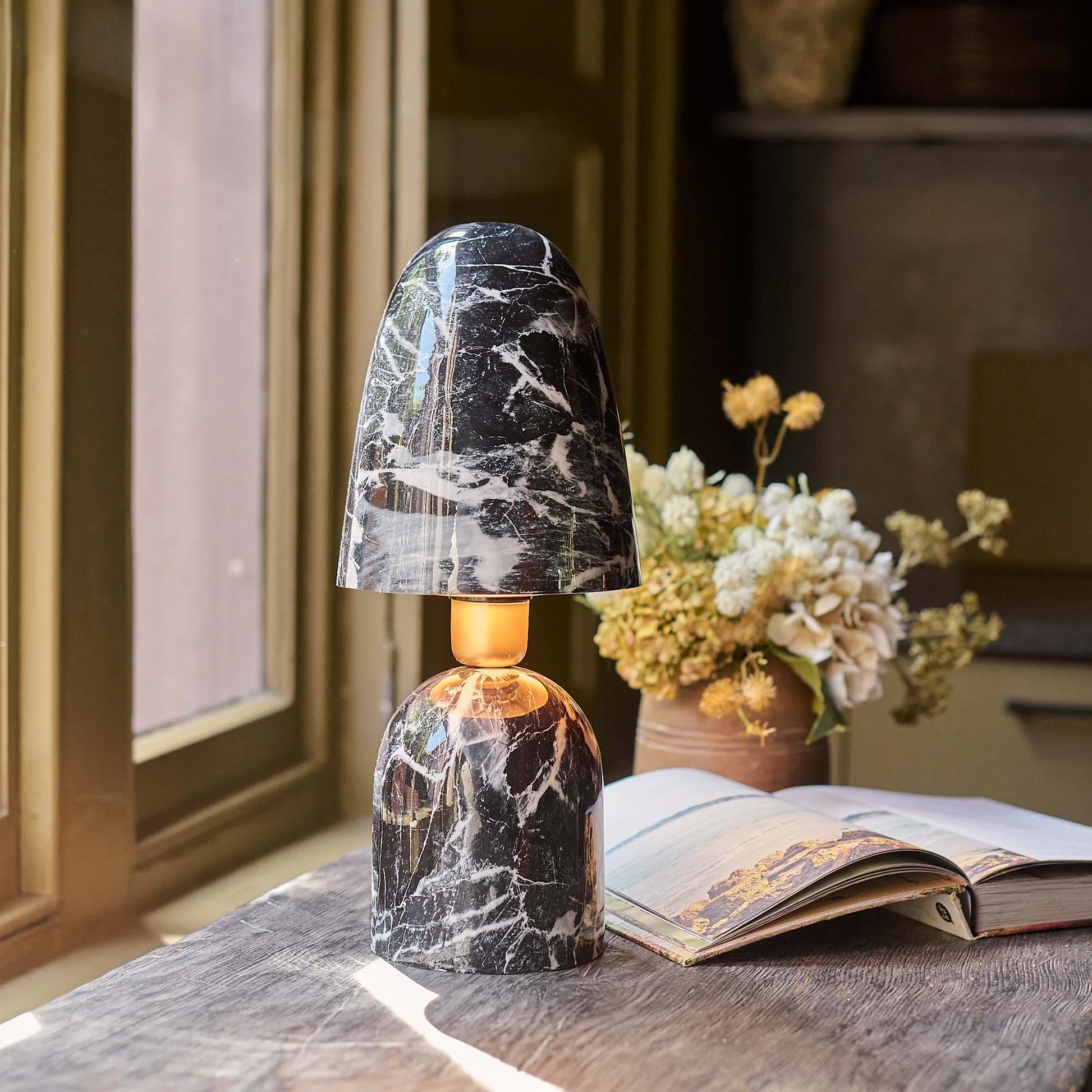 The Caswell Table Lamp by Abigail Ahern, featuring a black and white marble finish, sits on a wooden desk near an open book and dried flowers, providing stylish accent lighting beside a sunlit window.