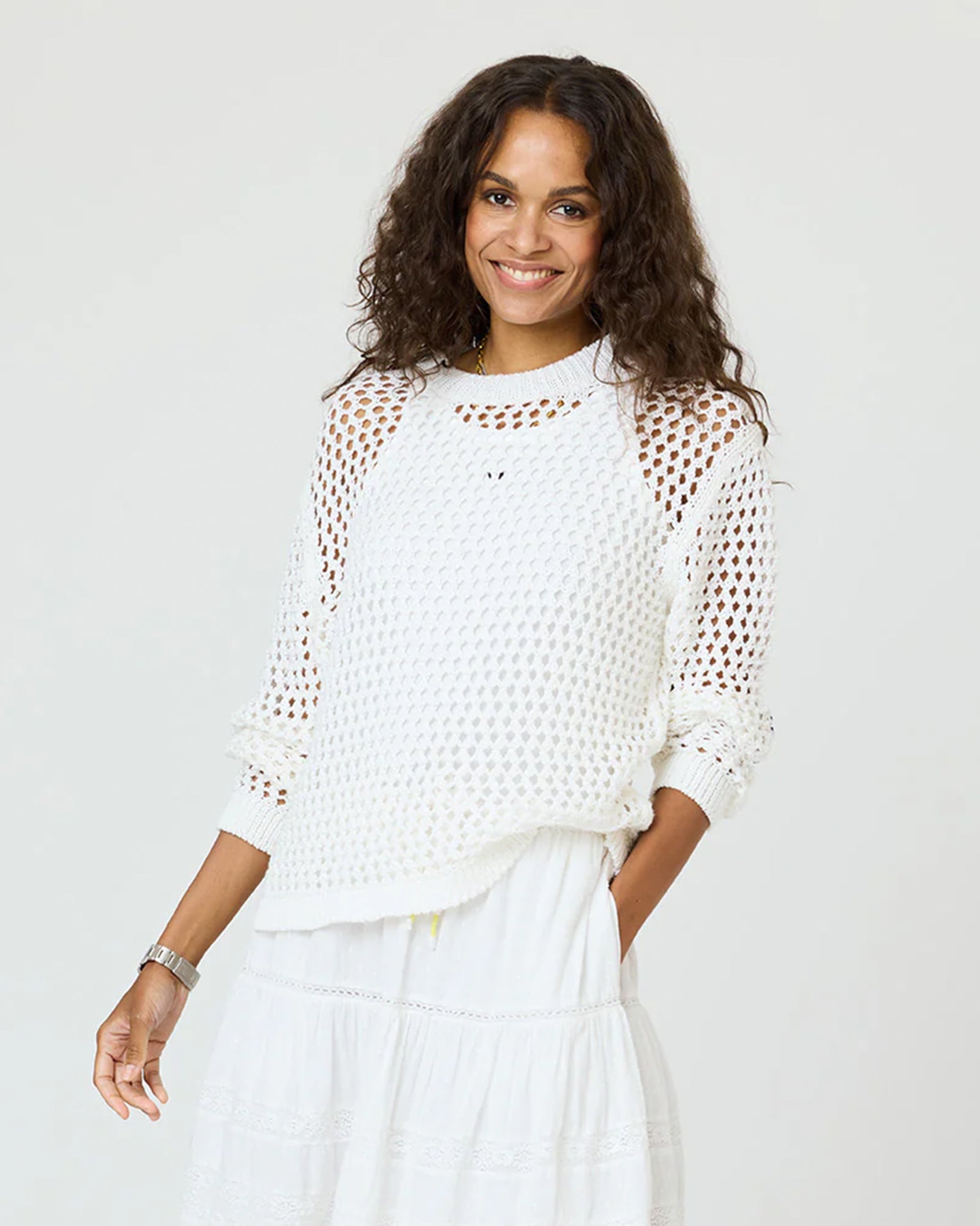 A woman with long curly hair smiles while wearing the Kerri Rosenthal Catch Open Stitch Sweater in white over a white dress, standing against a plain light background.